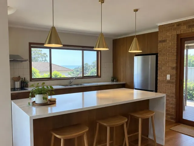 Modern kitchen with three brass pendant lights over a white island, wood cabinetry and brick wall in Curl Curl