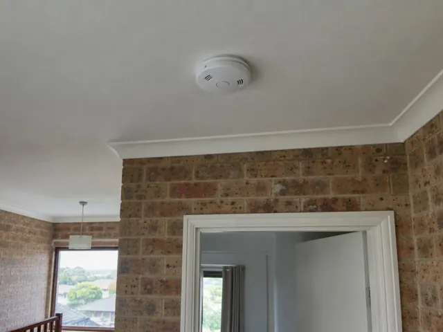 White smoke alarm mounted on a white ceiling above a brick wall interior in Curl Curl